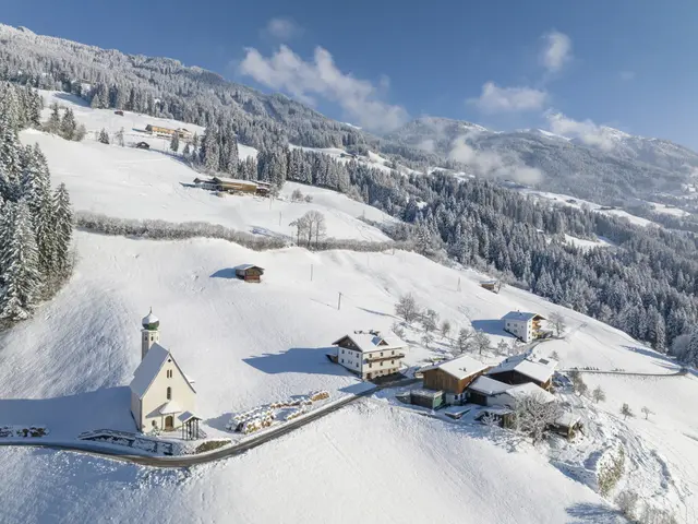 The image shows an aerial view of a small village nestled in the middle of a snowy mountain range....