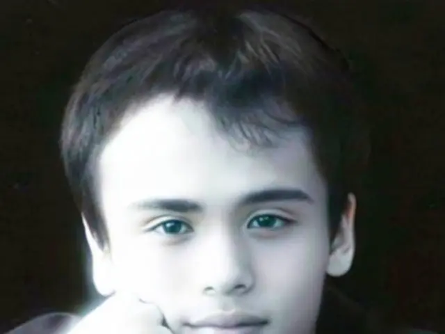 The image shows a young man in a black jacket posing for a picture against a dark background. He...