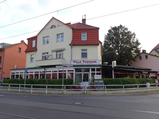 The image shows a road with a few vehicles driving on it, a railing, a few plants, and a few boards...