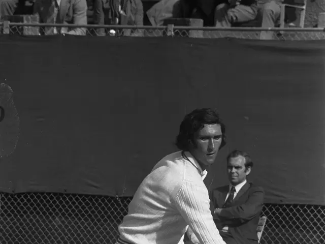 The image shows a man swinging a tennis racquet at a ball, with a mesh fencing in the background...