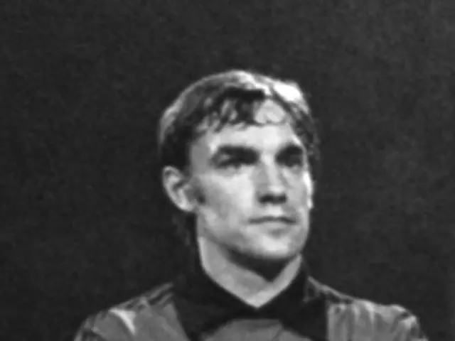 The image shows a black and white photo of a man standing on a soccer field with his arms crossed,...