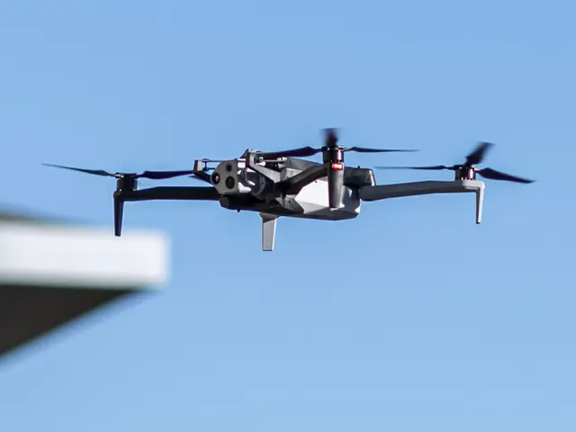 The image shows a drone flying in the air with a building on the left side and a clear blue sky in...