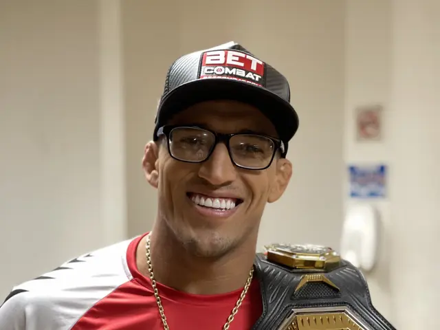 The image shows a man wearing a red and white t-shirt, a black cap, and glasses, with a gold chain...