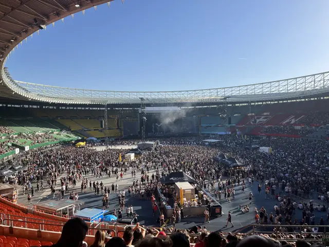 The image shows a large crowd of people gathered in a stadium, with a stage in the center. On the...