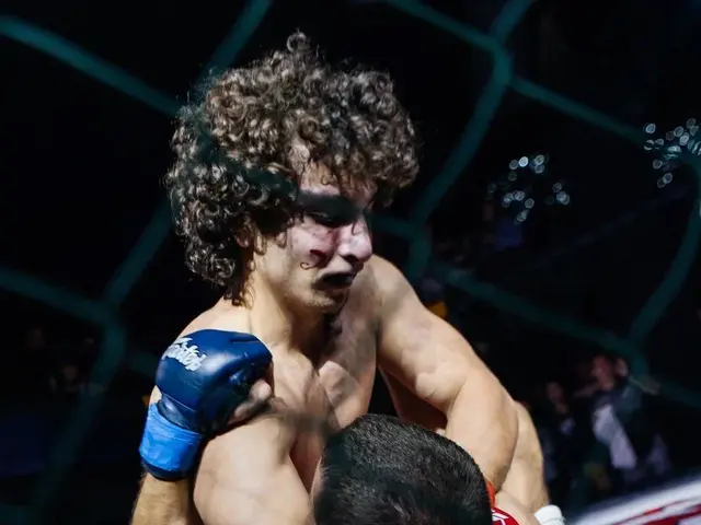 The image shows two men in a MMA ring, locked in a fierce battle. The ring is surrounded by a net,...