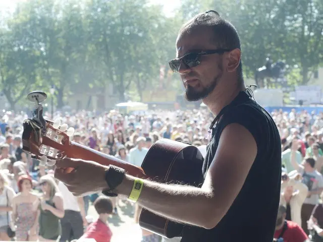 The image shows a man wearing sunglasses playing a guitar in front of a crowd of people, with trees...