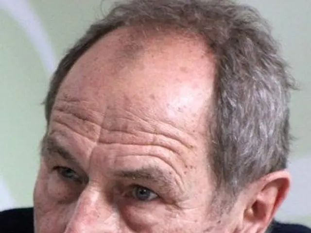 The image shows a close up of a man in a suit and tie, with a blurred background. He appears to be...