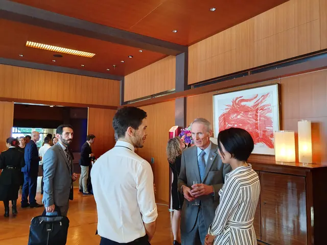 The image shows a group of people standing around talking to each other in a room with a wooden...