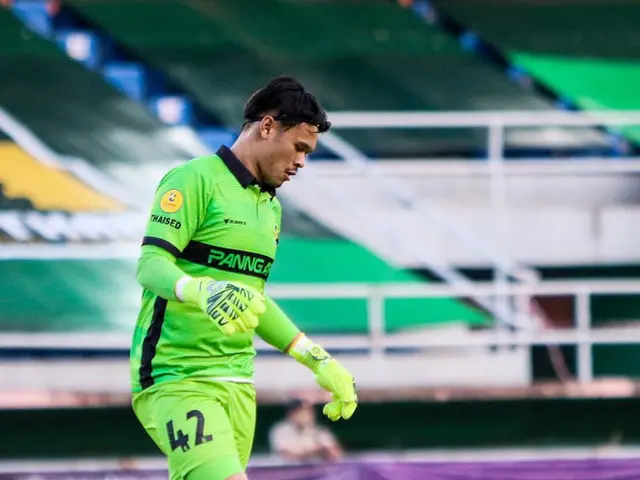 The image shows a man in a green goalkeeper's uniform kicking a soccer ball on a grassy field. In...