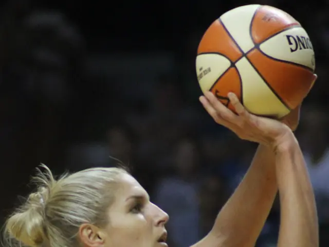 The image shows a woman holding a basketball in her hand, with a blurred background. She has a...