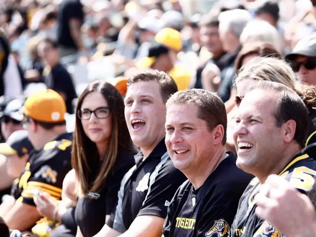 The image shows a group of people sitting in the stands at a football game, wearing black t-shirts...