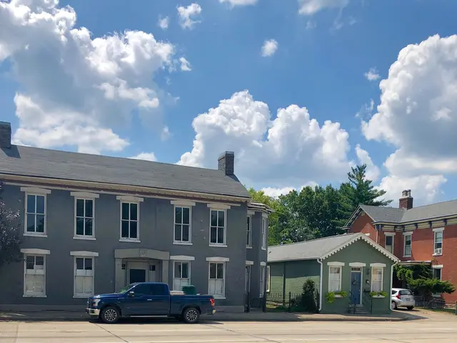 The image shows a street with cars parked on the side of it, buildings with windows, trees, and a...