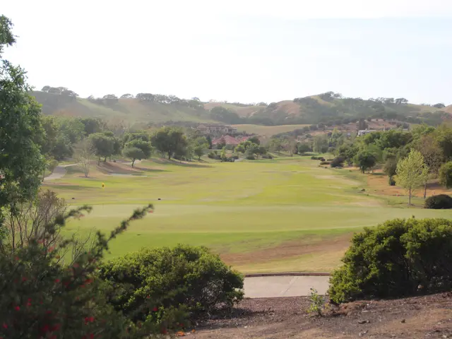 The image shows a golf course with lush green grass, tall trees, and various plants and flowers. In...