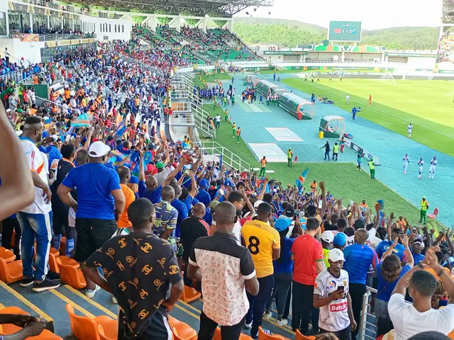 The image shows a large crowd of people standing in front of a soccer field, with some sitting on...