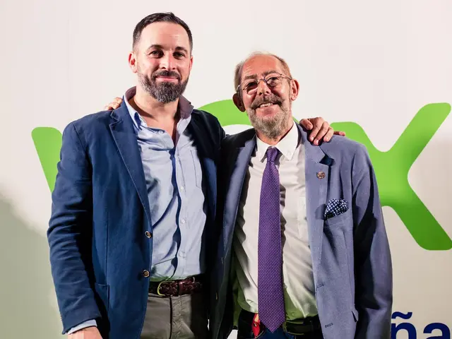 The image shows two men standing next to each other in front of a wall, both wearing suits and...
