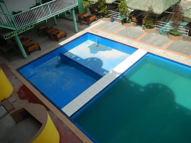 The image shows a swimming pool from the top of a building, surrounded by a railing, benches,...