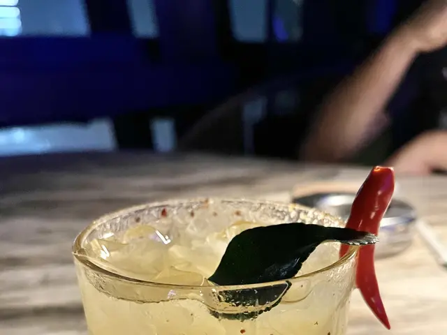 The image shows a glass filled with a drink sitting on top of a wooden table, with ice cubes, a...