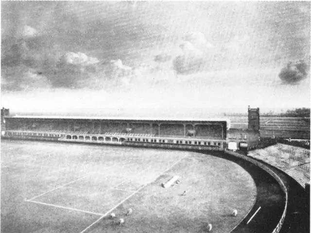 The image shows an old black and white photo of a soccer field with a stadium in the background....