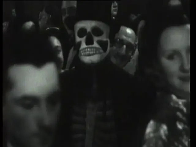 The image shows a group of people standing around each other in a dark room, with a man in the...