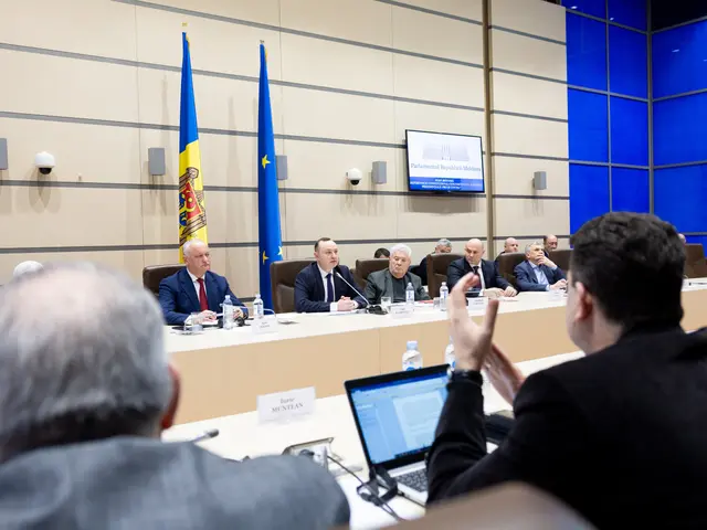 The image shows a group of people sitting around a table with laptops, bottles, name boards, mics...