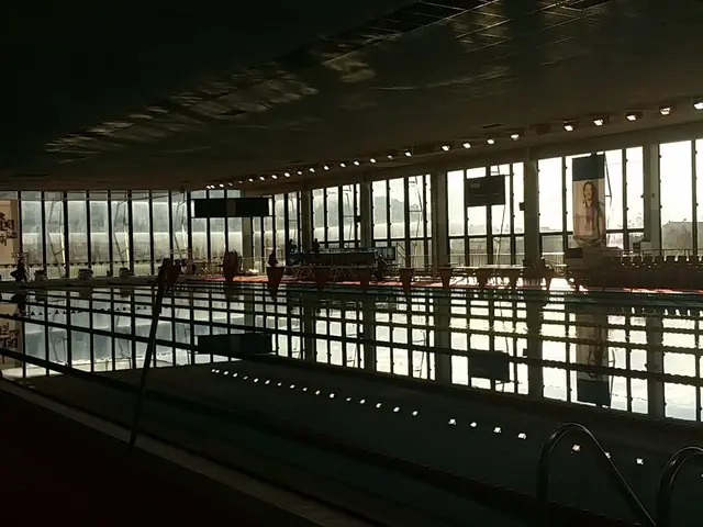 The image shows a large indoor swimming pool inside of a building, with a group of people standing...