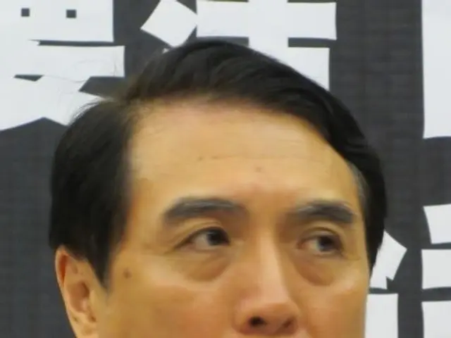 The image shows a man in a suit speaking into a microphone, with a banner in the background...