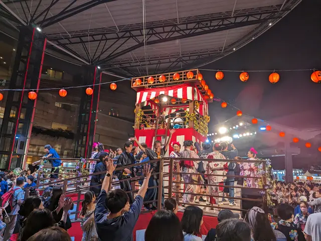The image shows a crowd of people standing on top of a stage in front of a building, illuminated by...