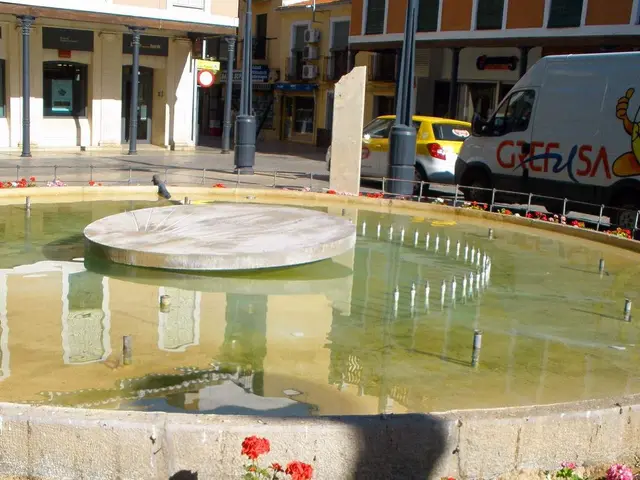 The image shows a small fountain in the middle of a city square surrounded by buildings with...