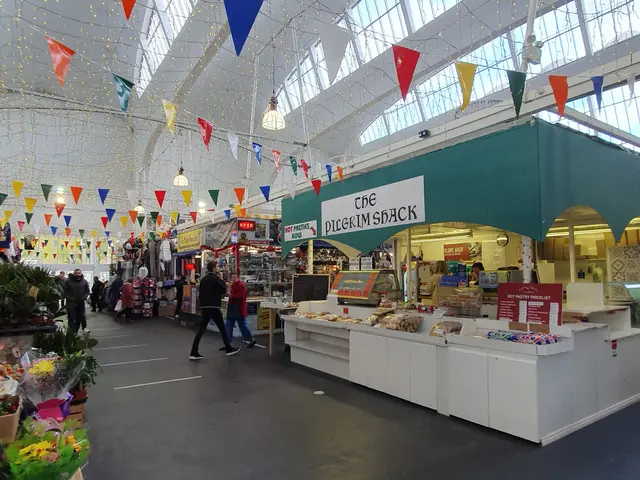 The image shows the inside of a market with lots of people walking around. On the left side of the...