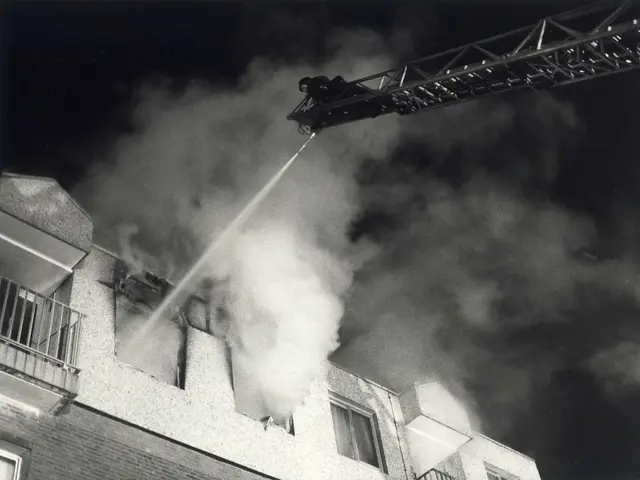 The image shows a black and white photo of a fire truck spraying water on a building with windows,...