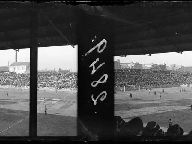 The image shows a black and white photo of a soccer game being played in a stadium, with a group of...