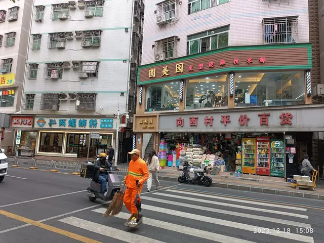 The image shows a man sweeping the street in front of a building with windows, air conditioners,...