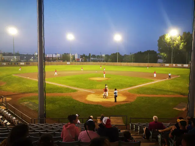 The image shows a baseball game being played in a stadium at night. There are people sitting in the...