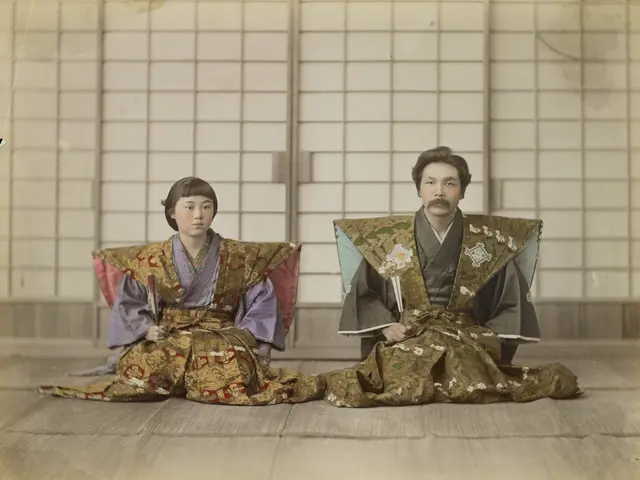 The image shows an old photo of a man and woman in kimono sitting on the floor in front of a wall....