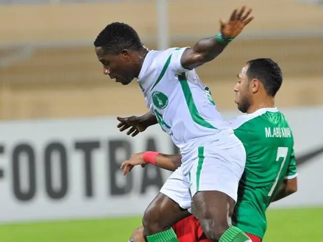 The image shows two men playing soccer on a grassy field, with a board in the background displaying...