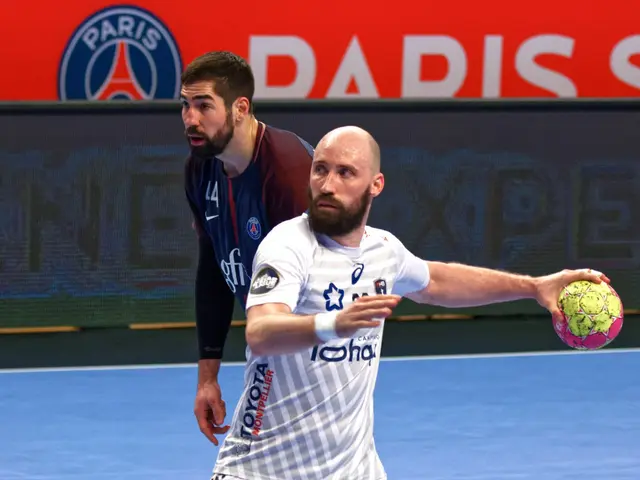 The image shows two men playing a game of handball on a court, with one of them holding a ball in...