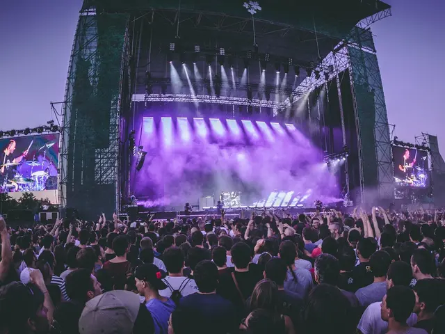 The image shows a large crowd of people gathered at a music festival, with a stage in the center of...