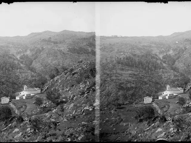 The image shows a black and white photo of a small village nestled in the mountains, surrounded by...