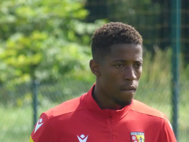 The image shows a young man in a red and yellow shirt standing on a soccer field surrounded by a...