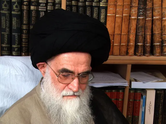 The image shows Ayatollah Ali Khamenei, the Supreme Leader of Iran, sitting on a chair and writing...