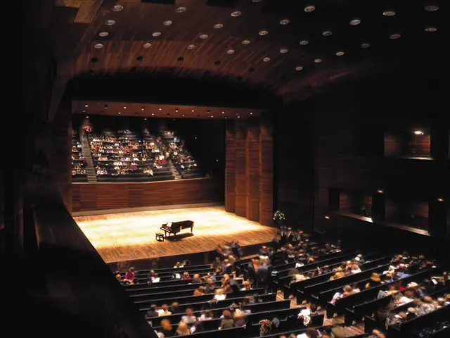 The image shows a large auditorium filled with lots of people sitting in chairs, with a piano on...