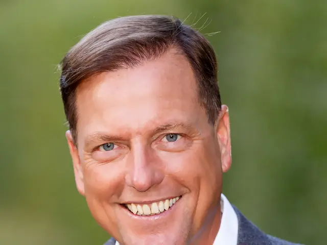 The image shows a man in a suit smiling at the camera with a blurred background. He appears to be...