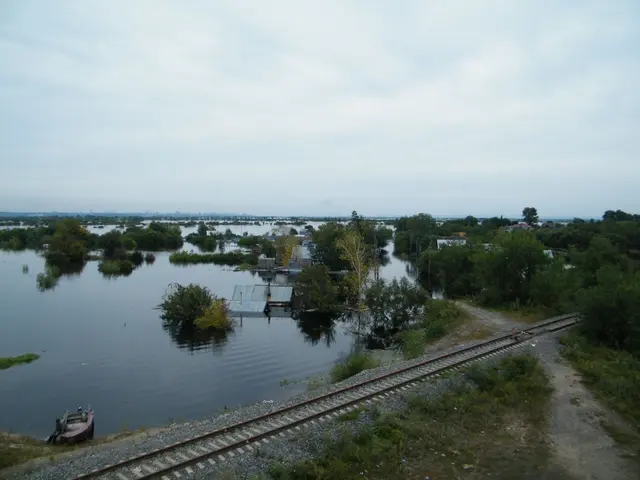 The image shows a train track running through a flooded area next to a body of water, with a boat...