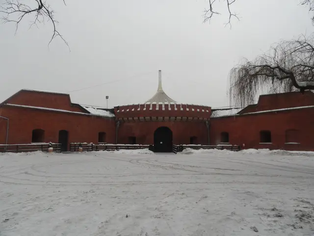 The image shows a large brick building with a dome on top of it, surrounded by a fence, with snow...