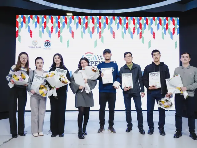 The image shows a group of people standing next to each other on a stage, each holding a bouquet of...