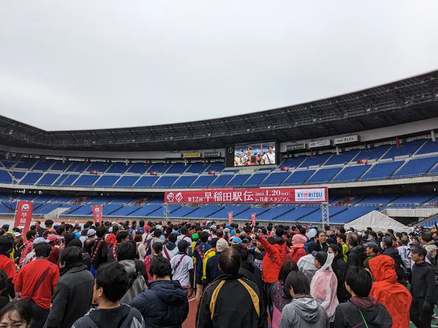 The image shows a large crowd of people standing in front of a stadium, with banners, chairs, and a...