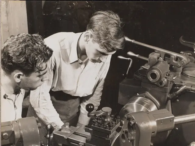 The image shows two men working on a machine in a factory. The image is in black and white and...