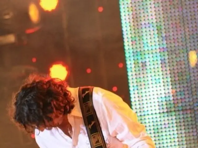 The image shows a man playing a guitar on stage at a concert, illuminated by bright lights. He is...