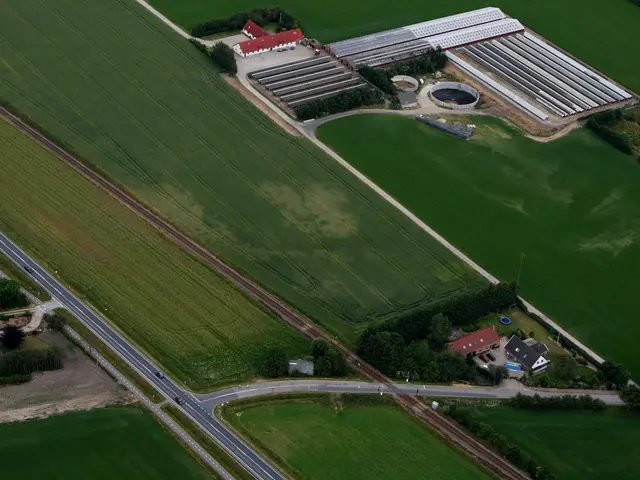 The image shows an aerial view of a farm in the middle of a green field, surrounded by trees,...