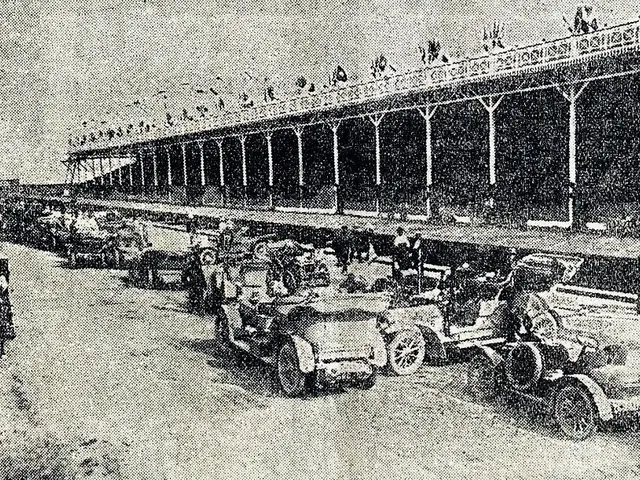 The image shows a black and white photo of a long line of cars parked in front of a building, with...